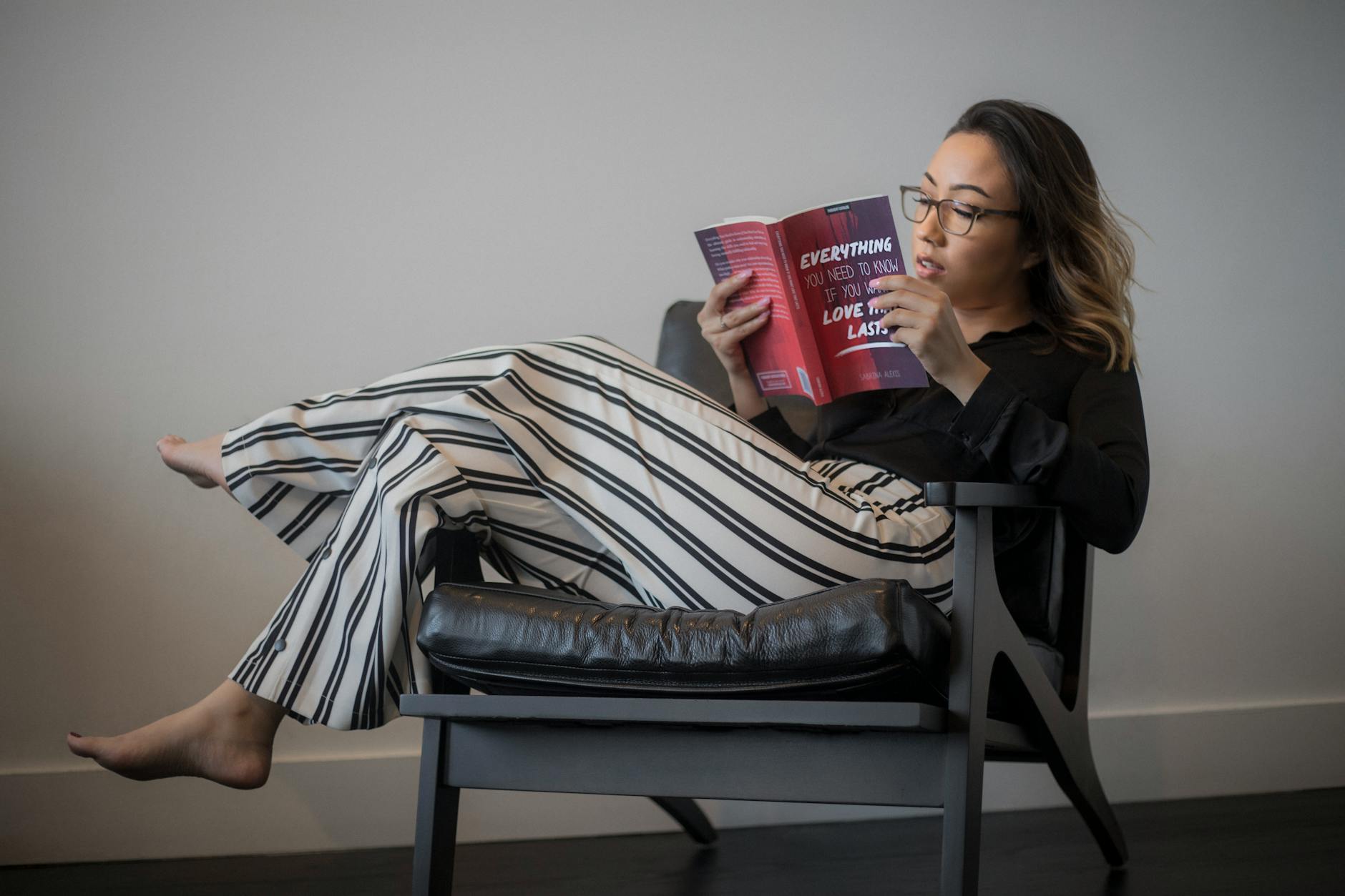 women s black long sleeved top white and black striped pants reading book sitting on gray wooden framed padded armchair